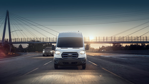 Ford E-Transit blanca en carretera al atardecer, con  puente al fondo.